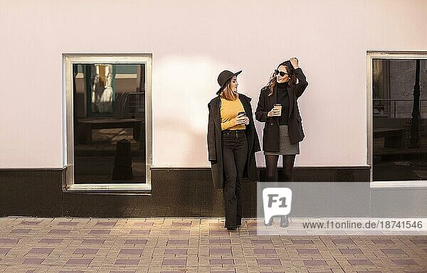 Zwei stilvolle Frauen Freundinnen in trendiger Kleidung genießen nettes Gespräch und trinken Kaffee zum Mitnehmen im Freien während eines Spaziergangs in der Stadt am Wochenende Tag. Weibliche Freundschaft Konzept