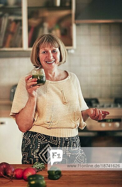 Glückliche Frau mittleren Alters mit frischem Gemüsesmoothie  die lächelt und in die Kamera schaut  während sie in der Küche zu Hause steht