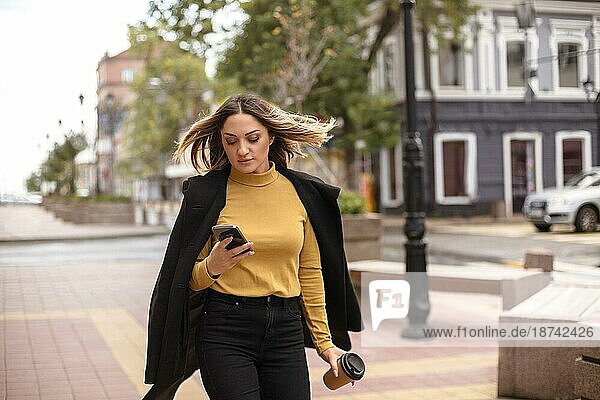 Junge attraktive Frau mit Smartphone zu Fuß Stadt Straße im Freien auf Herbst Tag  stilvolle Business Dame in schwarzem Mantel machen Telefonanruf auf dem Handy während des Spaziergangs außerhalb