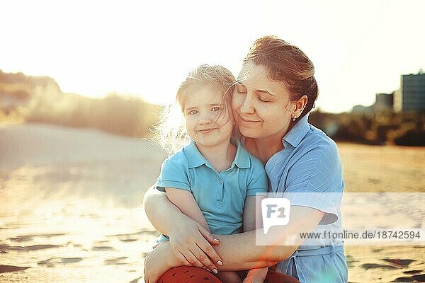 Junge liebende Mutter mit lächelnder Tochter  die ihre kleine Tochter am sonnigen Strand umarmt  glückliche Mutter umarmt kleines Mädchen und genießt die gemeinsame Zeit am Sommerstrand