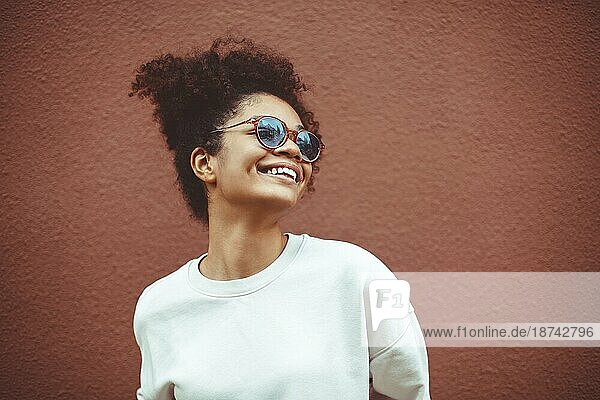 Junge Frau afrikanischer Abstammung mit stilvoller Sonnenbrille  mit lockigem Haar  das zu einem hohen Pferdeschwanz gebunden ist  schaut weg  während sie breit lächelt und gerade  perfekte Zähne zeigt  vor einem braunen Wandhintergrund posierend