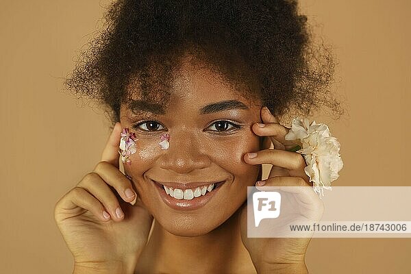 Porträt des schönen dunkelhäutigen Modells mit hoher Afrofrisur und natürlichem Make up mit Blütenblättern auf einer Seite ihres Gesichts hält rosa Nelke Blume in der Hand isoliert über beige Studio Wand Hintergrund