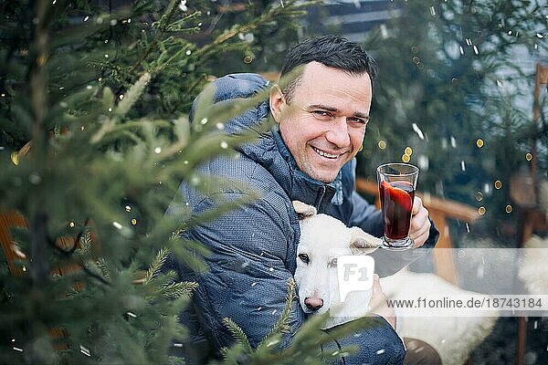 Lächelnder junger Mann sitzt auf einem Holzstuhl mit Hund auf dem Schoß  trinkt heißen aromatischen Grog  entspannter glücklicher Mann in warmer Kleidung  der sich im Hinterhof eines Privathauses während der Winterferien ausruht. Selektiver Fokus