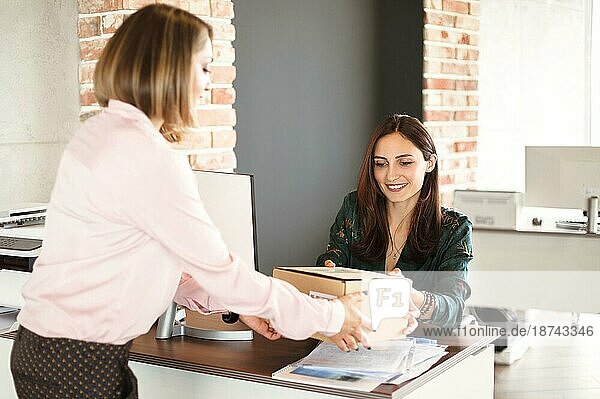 Fröhliche Managerin  die lächelt und einen Karton von einer Zustellerin entgegennimmt  während sie im Büro arbeitet