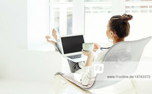 Barfüßige junge Frau mit Händen hinter dem Kopf  die auf einem Stuhl sitzt und sich mit geschlossenen Augen entspannt  nachdem sie einen Laptop in einem hellen Raum zu Hause benutzt hat