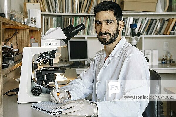 Junger Wissenschaftler vor dem Mikroskop  lächelt und schaut in die Kamera und notiert sich die Proben  die er im Labor untersucht