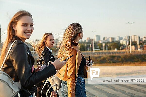 Glückliche junge Frauen mit Getränken zum Mitnehmen  die auf dem Bürgersteig spazieren gehen und an einem sonnigen Wochenendtag auf einer Straße in der Stadt lächeln