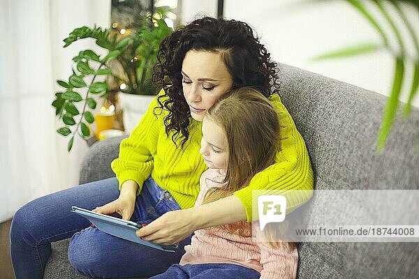 Mutter und kleines Mädchen Tochter beobachten Cartoons auf digitalen Tablet  während auf dem Sofa zu Hause sitzen  selektiven Fokus. Junge moderne Mutter und kleines Kind mit Gerät bei der Freizeitgestaltung zusammen