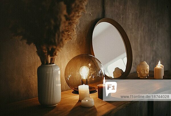 Runder Spiegel auf einem Holzregal mit zwei brennenden Kerzen und einem kleinen Porzellan Buddha Kopf  der sich in einer Ecke der verspiegelten Fläche auf einem Betonwand Hintergrund in einer minimalistischen modernen Wohnung spiegelt