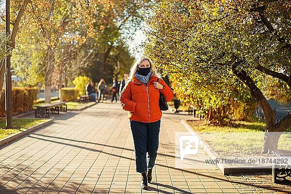 Ganzkörperfrau in Oberbekleidung und Maske  die auf einem gepflasterten Weg an einem sonnigen Tag im Herbstpark während einer Pandemie spazieren geht