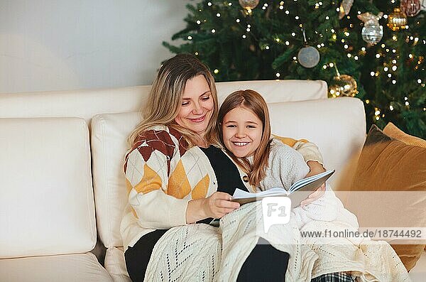 Glückliche Familie Großmutter und kleines Mädchen Tochter sitzen zusammen auf dem Sofa und Buch lesen  während Zeit zusammen zu Hause während der Weihnachtsferien im Winter  Umarmung und Abdeckung mit Wolle Plaid