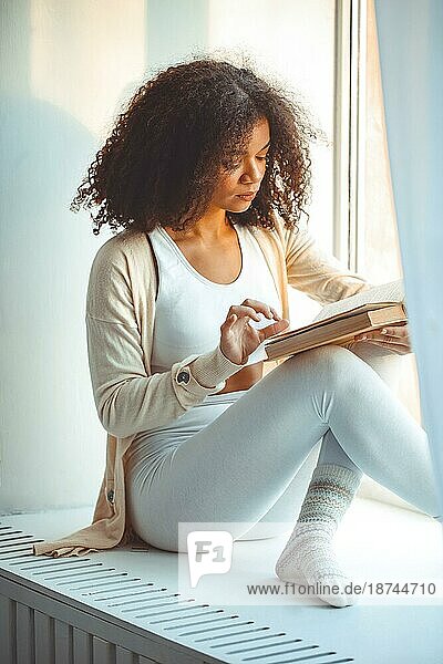 Mein Hobby ist das Lesen. Ruhig entspannt afrikanische amerikanische junge Frau  die Buch liest  während sie auf der Fensterbank zu Hause sitzt  schwarzes lockiges Mädchen am Fenster  das sich entspannt und seine Freizeit am Wochenendtag verbringt  vertikaler Schuss