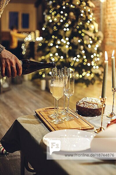 Silvesterfeier. Mann gießt Champagner in Gläser stehen auf dem Tisch mit festlichen Weihnachtsessen  Kerzen und eingepackt Geschenke gegen unscharfen Hintergrund von Backstein Kamin und firtree  beschnitten