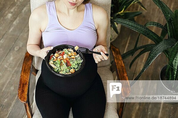 Schwangere junge brünette Frau ißt gesunden Salat aus Schüssel Draufsicht. Gesundes Essen beim Mittagessen auf einem gemütlichen Sessel