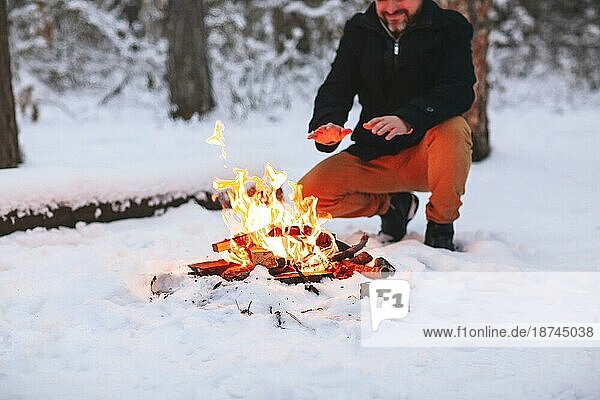 Reifer Mann wärmt sich die Hände am Feuer  nachdem er mitten im verschneiten Wald am kalten Winterabend ein Lagerfeuer gemacht hat  er nutzt die Wärme  um sich bei schlechtem Wetter wohl zu fühlen und genießt den Winterurlaub