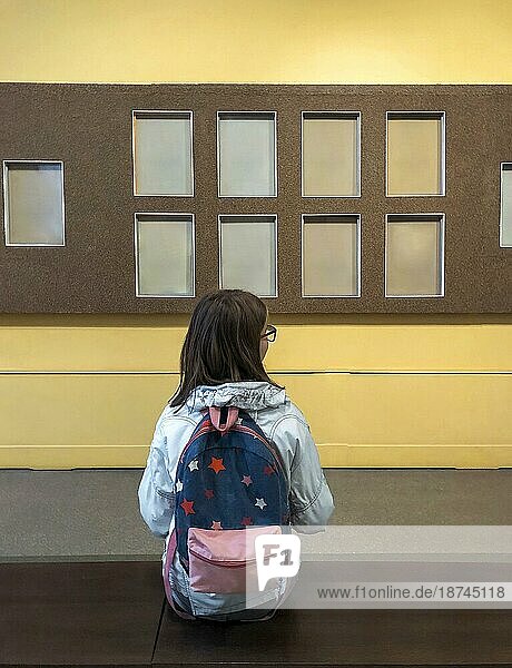 Rückenansicht eines Mädchens mit Rucksack in einem Museum  das eine Ausstellung besucht  eine Schülerin  die auf einer Bank in einer Kunstgalerie sitzt und mit Interesse Kunstobjekte oder Exponate an der Wand betrachtet