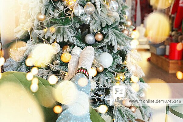 Weihnachten und Neujahr Hintergrund mit weiblichen Füssen in warmen gestrickten Socken gegen Weihnachtsbaum mit gelben Glühbirnen Girlande geschmückt  selektiven Fokus. Frau entspannt zu Hause in der Weihnachtszeit  beschnittenes Foto