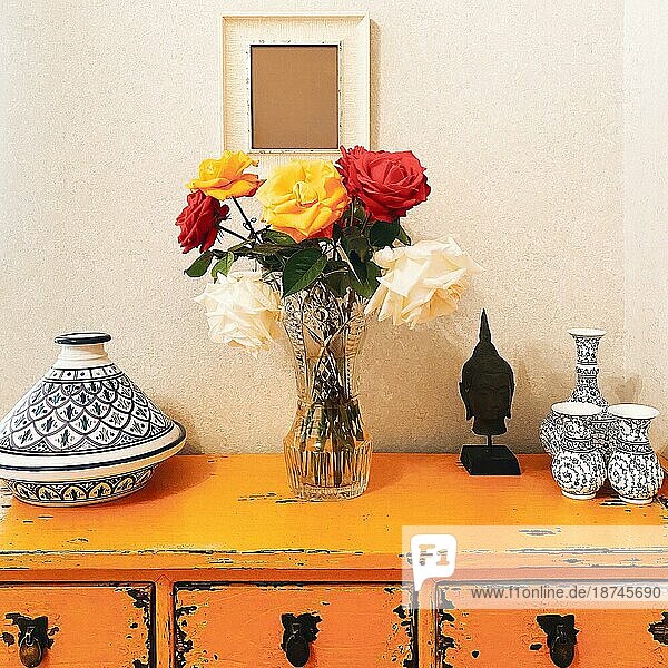 Schöner Strauß frischer mehrfarbiger Rosenblüten in Glasvase auf gelbem Vintage Tisch mit Buddhakopf und verschieden großen antiken Keramikvasen  ohne Menschen. Warm und gemütlich nach Hause Dekor