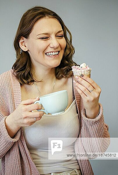 Glückliche junge Frau ißt leckeren rosa Muffin mit einem breiten Lächeln und geschlossenen Augen  nachdem sie einen Bissen genommen und probiert hat. Sie hält eine Tasse mit einem heißen Getränk in der Hand  vorgrauem Hintergrund. Ungesundes Essen Konzept