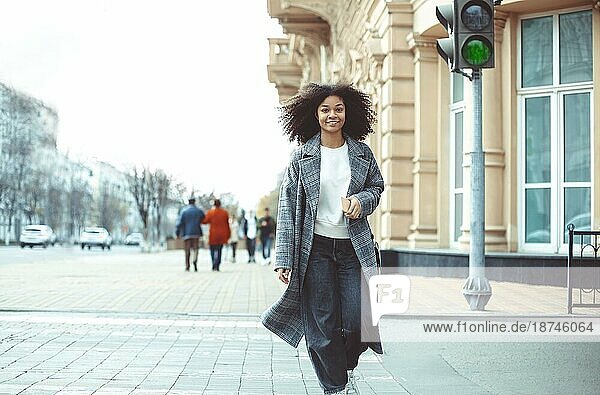 Junge stilvolle positive afroamerikanische Frau überqueren Zebrastreifen und lächelnd in die Kamera beim Gehen in der Stadt im Herbst Tag. Glückliches schwarzes Mädchen auf dem Zebrastreifen im Freien. Menschen auf den Straßen der Stadt
