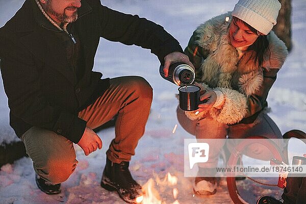 Joyful glückliche Familie Paar in der Liebe Sonnenbaden am Lagerfeuer draußen im Winter verschneiten Wald trinken heißen Tee zusammen  Mann und Frau wärmen sich umarmen und liebevollen Blick auf einander. Trekking  Abenteuer und saisonalen Urlaub