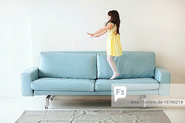 Kleine kaukasische niedlich schönes Mädchen in gelben Kleid spielen und springen auf blaün Sofa im Wohnzimmer zu Hause vor weißem Hintergrundem Betonwand Hintergrund. Kindheit und Freizeit Konzept