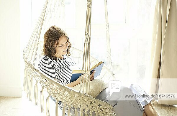 Ganzer Körper erfreut junge Frau ruht in Hängesessel und Lesen Buch am Wochenende Tag in sonnenbeschienenen Raum zu Hause