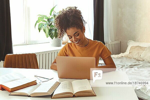 Fröhliche junge afroamerikanische Studentin  die zu Hause am Schreibtisch sitzt  umgeben von Lehrbüchern  vor einem Laptop  der fleißig studiert und sich auf Prüfungen im Internet vorbereitet  und mit einem zufriedenen Lächeln in den Laptop schaut