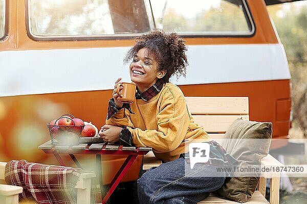 Junge afroamerikanische Frau sitzt am Tisch in der Nähe von Auto im Herbst Wald und trinken heißen Tee  verträumt gemischter Rasse weiblichen entspannenden rund um Campingplatz  Zeit in der Natur. Camping  Reisen und Vanlife