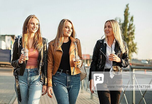 Glückliche junge Frauen mit Getränken zum Mitnehmen  die auf dem Bürgersteig spazieren gehen und an einem sonnigen Wochenendtag in der Stadt lächeln