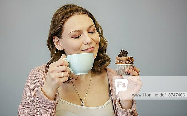 Glückliche junge Frau ißt leckeren rosa Muffin mit geschlossenen Augen  nachdem sie einen Bissen genommen und probiert hat  und hält eine Tasse mit heißem Getränk  vorgrauem Hintergrund. Ungesundes Essen Konzept