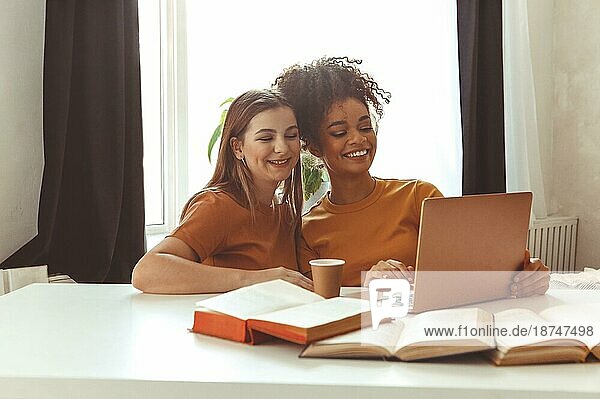 Zwei glückliche Freundinnen  die sich auf Prüfungen vorbereiten  sitzen zusammen vor einem Laptop neben aufgeschlagenen Lehrbüchern vor dem Hintergrund eines Wohnzimmerinterieurs  fröhliche Klassenkameraden  Studenten  die gemeinsam im Haus lernen