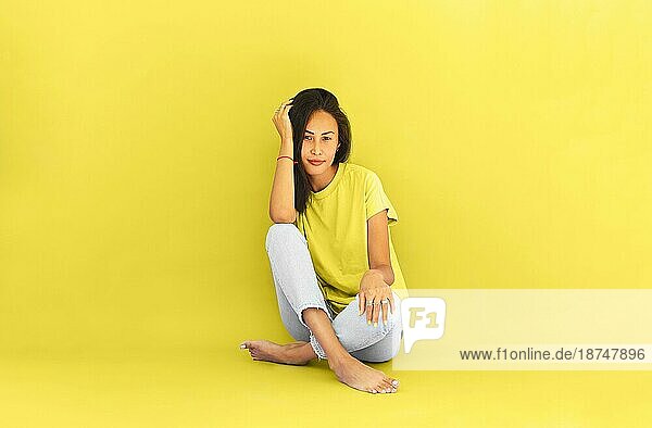 Ganzkörperdarstellung einer asiatischen Frau in Freizeithemd und Jeans  die barfuß auf dem Boden in einem hell erleuchteten gelben Studio sitzt