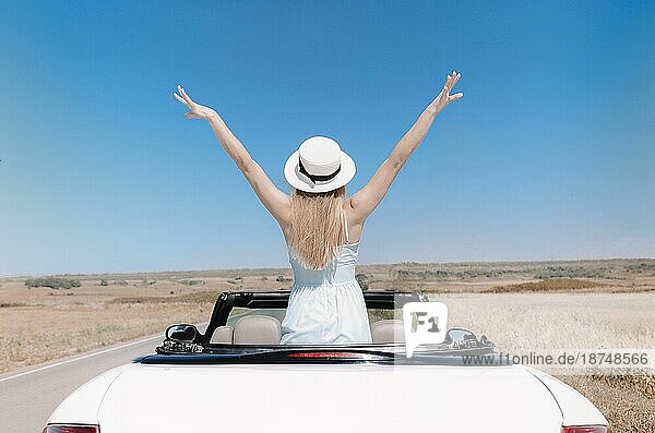 Rückenansicht einer fröhlichen jungen Frau in Sommerkleidung im Cabrio während einer Autofahrt