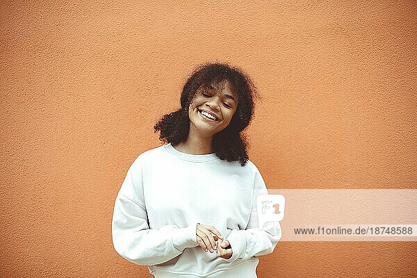Pures Glück. Nette glückliche afroamerikanische Mädchen mit lockigen Frisur trägt weißen Pullover posiert mit geschlossenen Augen gegen orange Wand. Lächelnde schwarze junge Frau  die mit ihrem Leben zufrieden ist