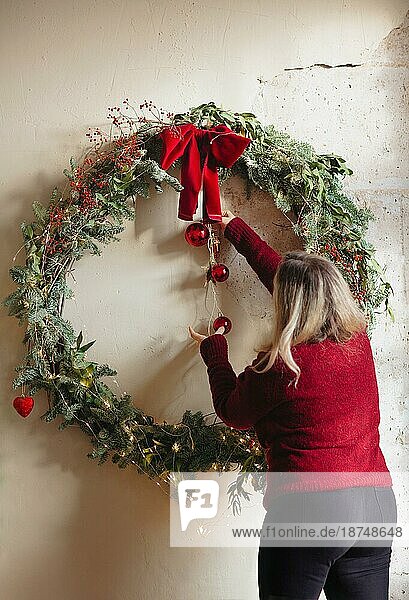 Seitenansicht einer erwachsenen Frau in einem roten Pullover  die am Weihnachtstag zu Hause Dekorationen an die Zweige eines Nadelbaums hängt
