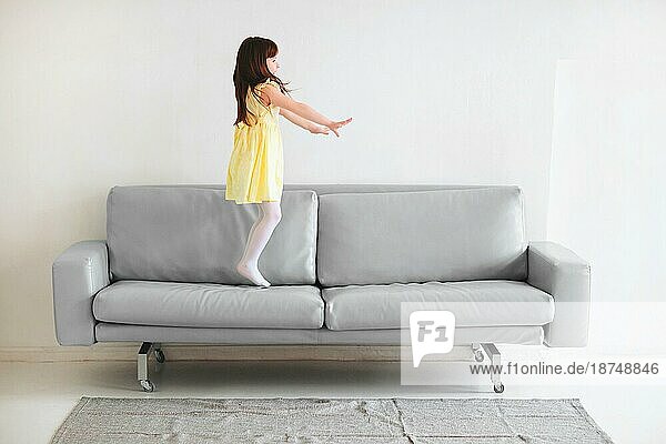 Kleine kaukasische niedlich schönes Mädchen in gelben Kleid spielen und springen auf grauen Sofa im Wohnzimmer zu Hause vor weißem Hintergrundem Betonwand Hintergrund. Kindheit und Freizeit Konzept