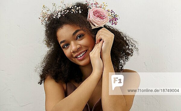Close up Frühling Porträt der wunderschönen lächelnden afrikanisch amerikanischen jungen Frau mit frischen rosa Rose Blume in lockiges Haar Blick in die Kamera  vorgrauem Hintergrund. Natürliche Schönheit der afrikanischen Frauen
