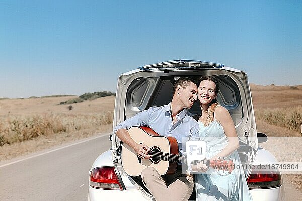 Glückliches junges Paar spielt Gitarre und genießt die gemeinsame Zeit  während es auf dem offenen Kofferraum eines auf einer Landstraße geparkten Autos sitzt