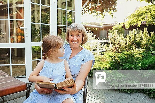 Ältere Großmutter und Enkelin verbringen Zeit zusammen auf der sonnigen Terrasse in der Nähe von Haus  Oma liest Märchen laut zu reizenden Mädchen  während kleines Mädchen sitzt auf ihrem Schoß Sommerurlaub mit Großeltern