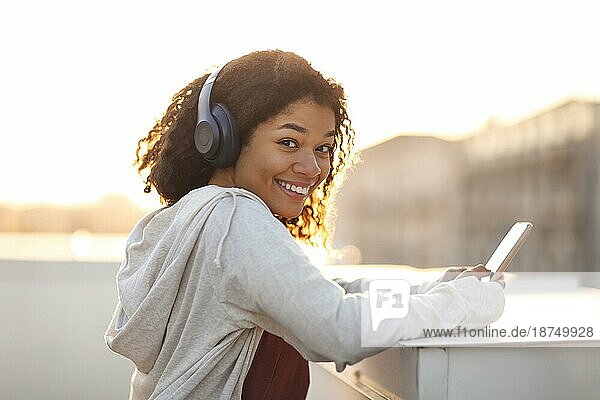 Seitenansicht Porträt der glücklichen fröhliche afrikanische amerikanische Frau in Kopfhörer hören Musik auf dem Smartphone und lächelnd in die Kamera während der Morgengymnastik auf dem Dach