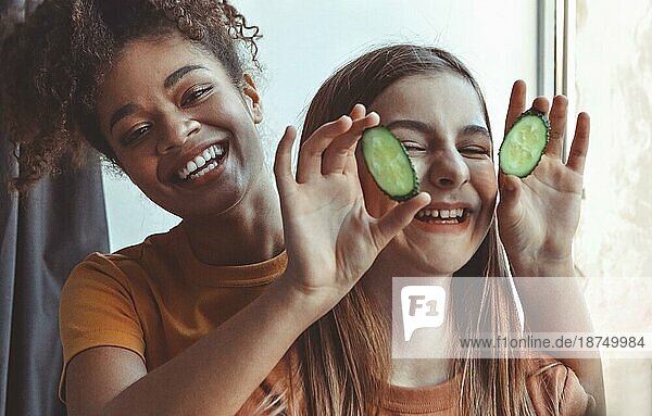 Close up über Foto von zwei besten Freundinnen verbringen Zeit zusammen Spaß beim Auftragen von Gurkenscheiben auf die Augen halten sie mit den Händen  Blick in die Kamera breit lächelnd. Freundschaft Konzept
