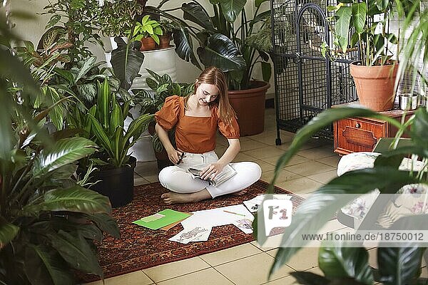 Junge rothaarige Frau  die ihre Feng Shui Wunschkarten auf dem Teppich im Zimmer im Boho Stil mit vielen Pflanzen anlegt. Träume und Wünsche