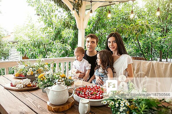 Liebevolle Familie mit Kindern am Tisch sitzend mit leckeren Desserts und Blumen beim Frühstück auf dem Land im Sommer
