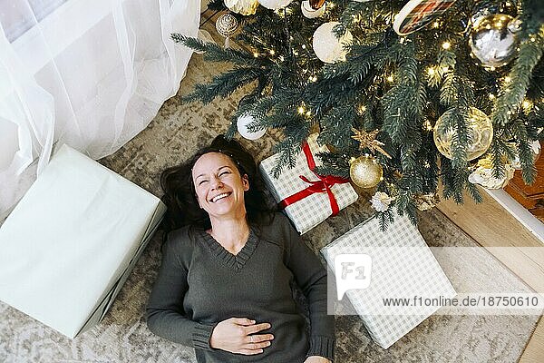 Glückliche junge kaukasische Frau  die unter einem geschmückten Weihnachtsbaum auf dem Boden liegt. Lächelndes Mädchen in einem gestrickten Pullover  das das neue Jahr feiert. Glücklich und Urlaub Konzept