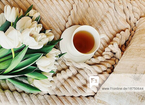 Strauß frischer weißer Tulpen und weiße Tasse Kaffee auf gemütlichem Bett mit beiger Plüschdecke und Kissen in hellem Schlafzimmer platziert