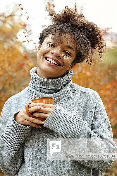 Junge glückliche lächelnde gemischtrassige Frau mit Kaffeetasse in herbstlicher Natur  zufriedene Afroamerikanerin mit lockigem Haar im Strickpullover  die in die Kamera schaut  während sie den Morgen in einem schönen Herbstgarten genießt