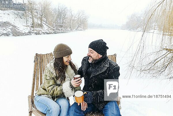 Glückliches  hübsches Paar mittleren Alters in warmer Kleidung  das während eines romantischen Wintertreffens in der Natur heißen Tee trinkt  ein verliebter Mann und eine verliebte Frau  die auf einem zugefrorenen See unter fallendem Schnee sitzen und sich an einem kalten  frostigen Tag unterhalten