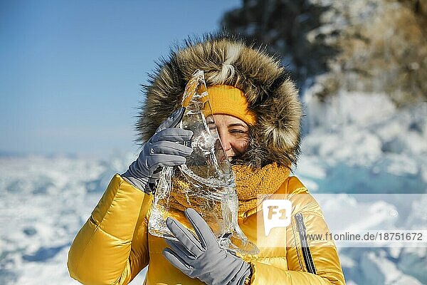 Glücklich lächelnde Frau in gelber Winterjacke  die ein Stück durchsichtiges  zerkleinertes Eis über ihr Gesicht hält. Die Sonne scheint durch die Seiten der Eiswürfel. Die Eiswürfel sehen aus wie Diamanten