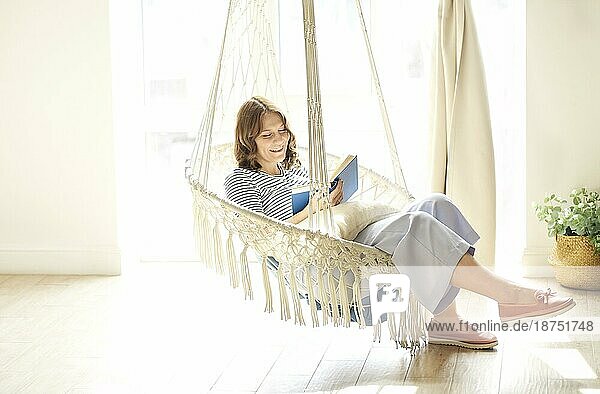 Ganzer Körper erfreut junge Frau ruht in Hängesessel und Lesen Buch am Wochenende Tag in sonnenbeschienenen Raum zu Hause